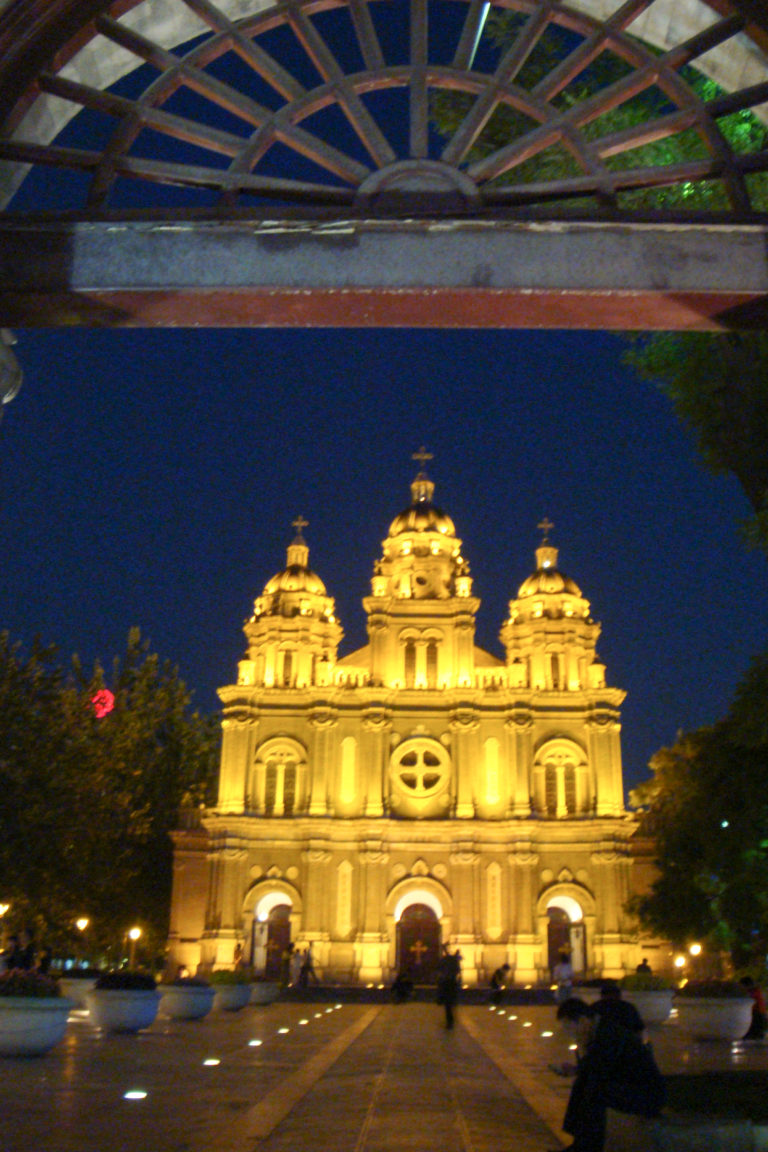 Nighttime exterior view of St. Joseph's Church on Wangfujing in Beijing, with its three-tower Baroque-influenced facade illuminated against a dark blue sky.