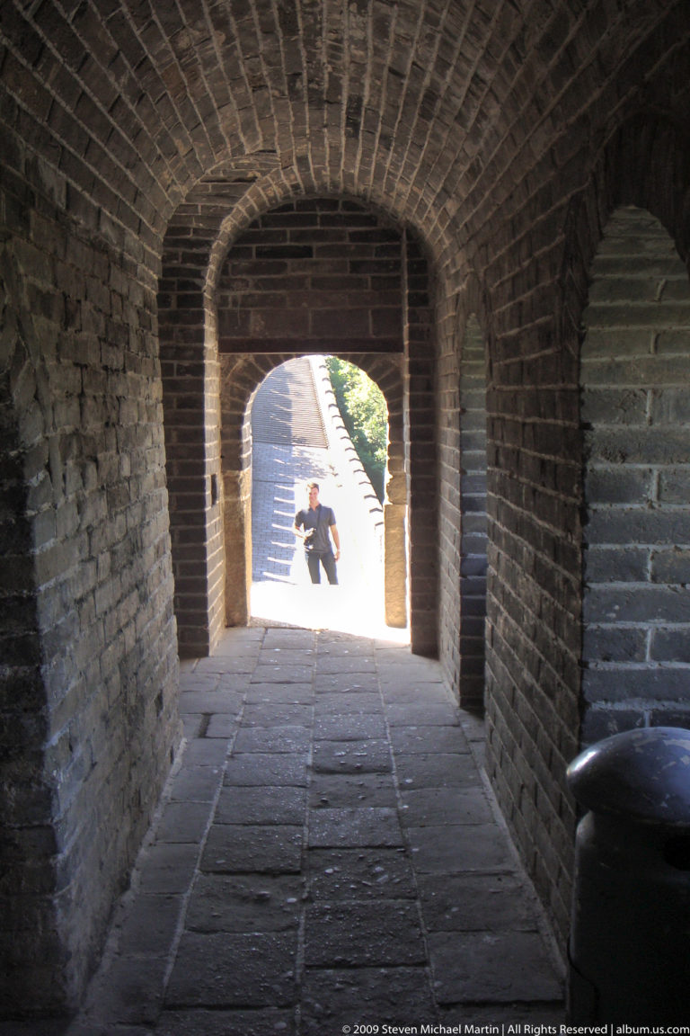 Interior of a Great Wall watchtower at Mutianyu, looking through a deep brick arched doorway to a sunlit rampart where a visitor stands.