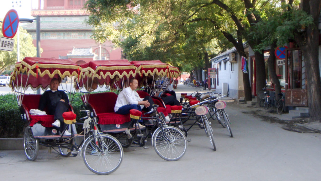 Several pedicab drivers wait in a line of red-canopied cycle rickshaws on a narrow, tree-lined Beijing hutong street.