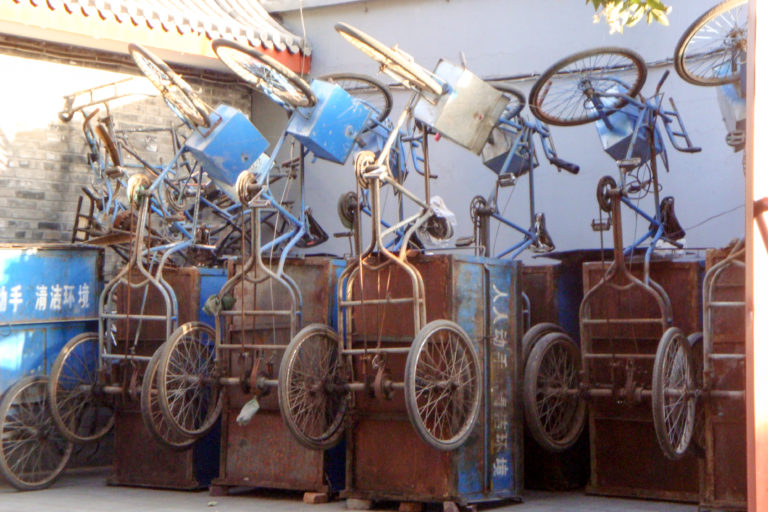 Rusty three-wheeled cargo tricycles stored upright on their metal boxes against a wall in a Beijing hutong.