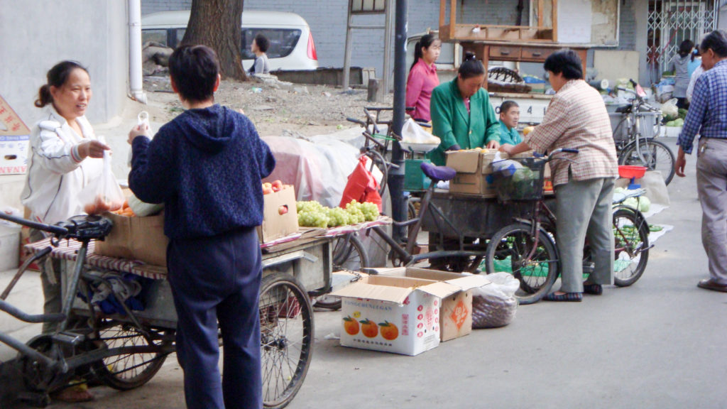 Women selling and sorting vegetables from loaded cargo tricycles in a narrow Beijing hutong street.