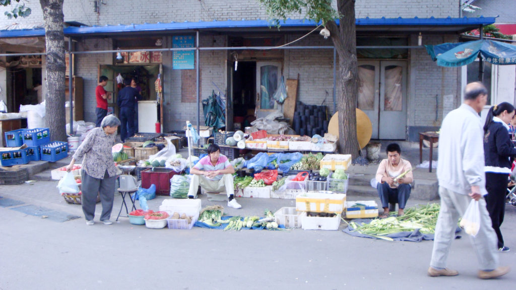 Informal curbside produce stall in a Beijing hutong, with vendors selling vegetables, fruit, eggs, and household goods outside a gray-brick storefront.