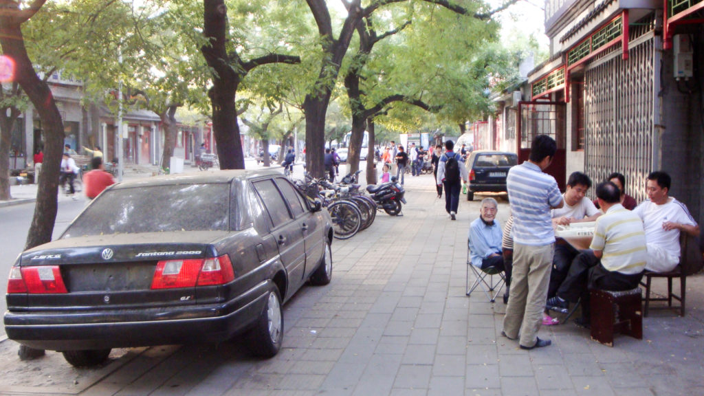 Wide view of a tree-lined Beijing hutong sidewalk where residents sit at outdoor tables eating and talking beside parked bicycles, scooters, and a black sedan.