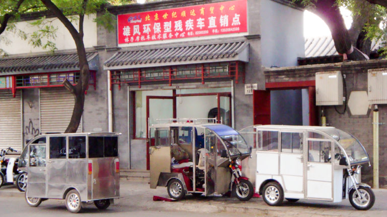 Several enclosed three-wheeled vehicles parked in front of a gray-brick hutong storefront with a red Chinese shop sign above.