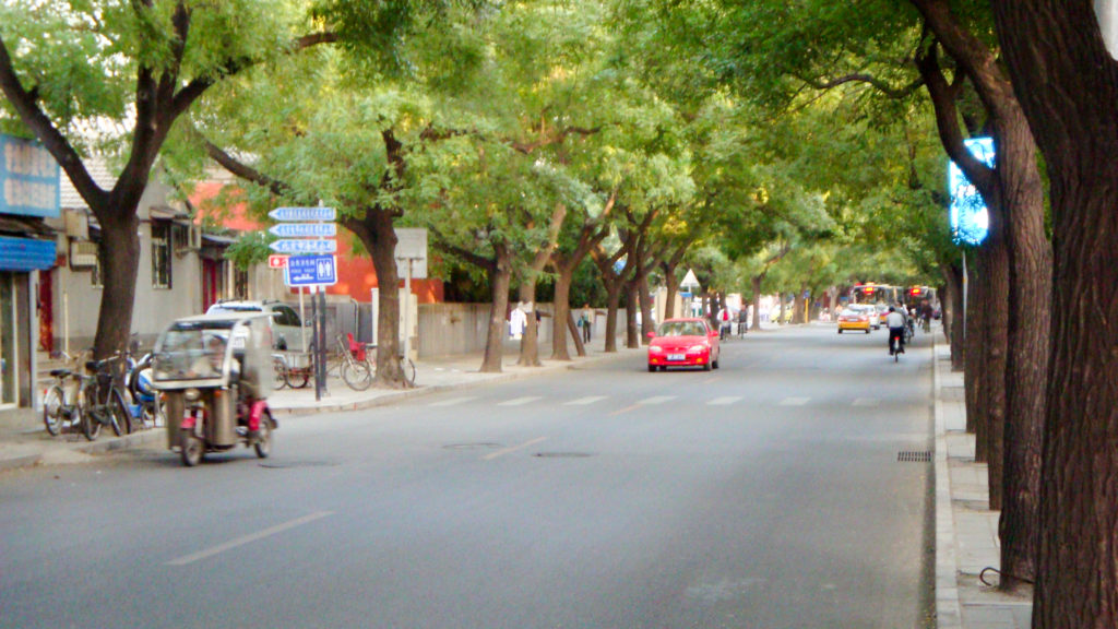 Tree-lined neighborhood street at the edge of a Beijing hutong district with low-rise buildings and mixed car and bicycle traffic.