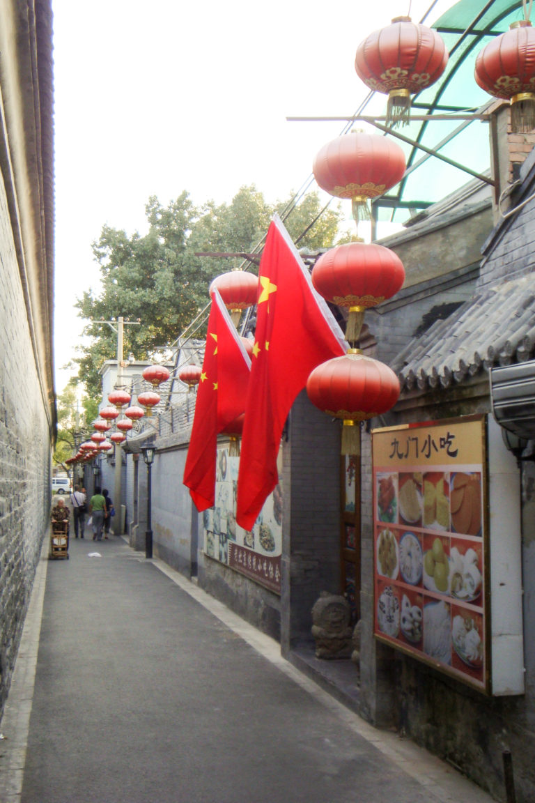 Wide view down a gray-brick Beijing hutong lined with Chinese flags, red lanterns, and a 九门小吃 snack-shop sign.