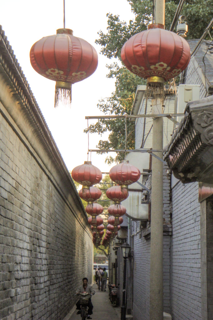 Narrow gray-brick hutong alley in Beijing with high walls and rows of red lanterns hanging overhead.