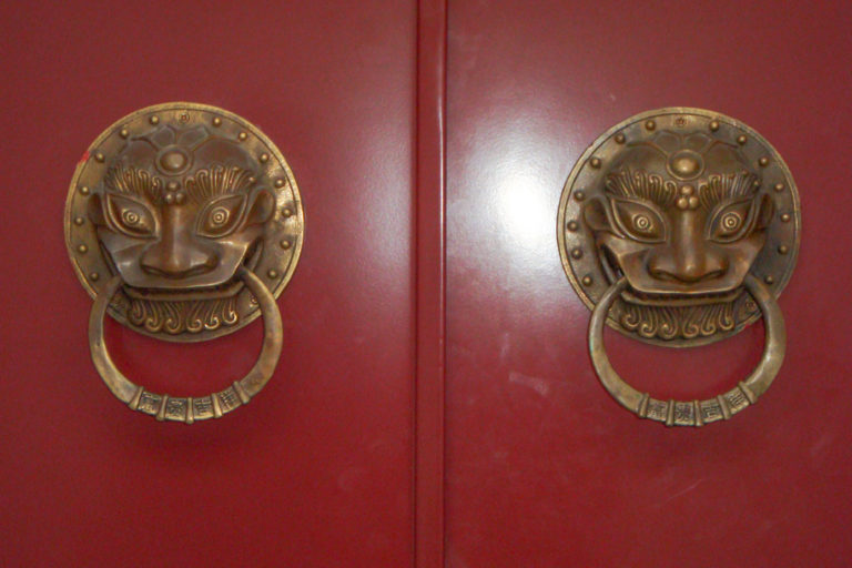 Close-up of two symmetrical lion-mask brass door knockers on a glossy red double wooden door.