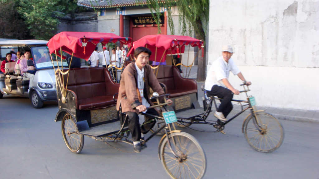 Red-canopied cycle rickshaw and a bicyclist riding side by side on a gray-walled Beijing hutong street with a traditional gate in the background.