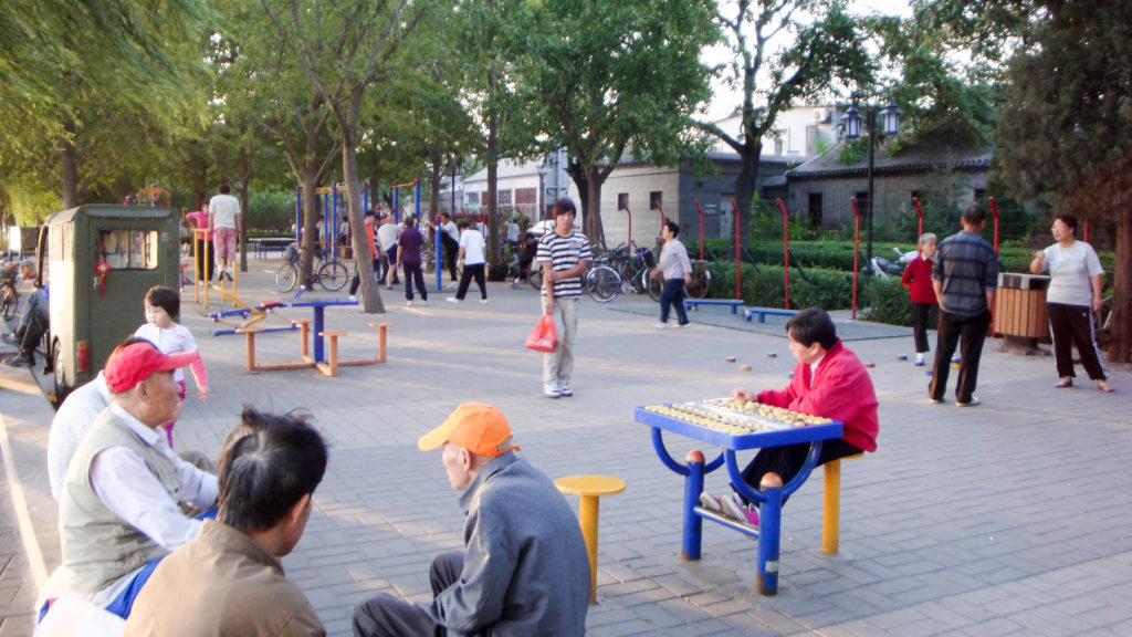 Older residents in a Beijing hutong courtyard sit at tables playing a board game while others walk and use outdoor exercise equipment under mature trees.