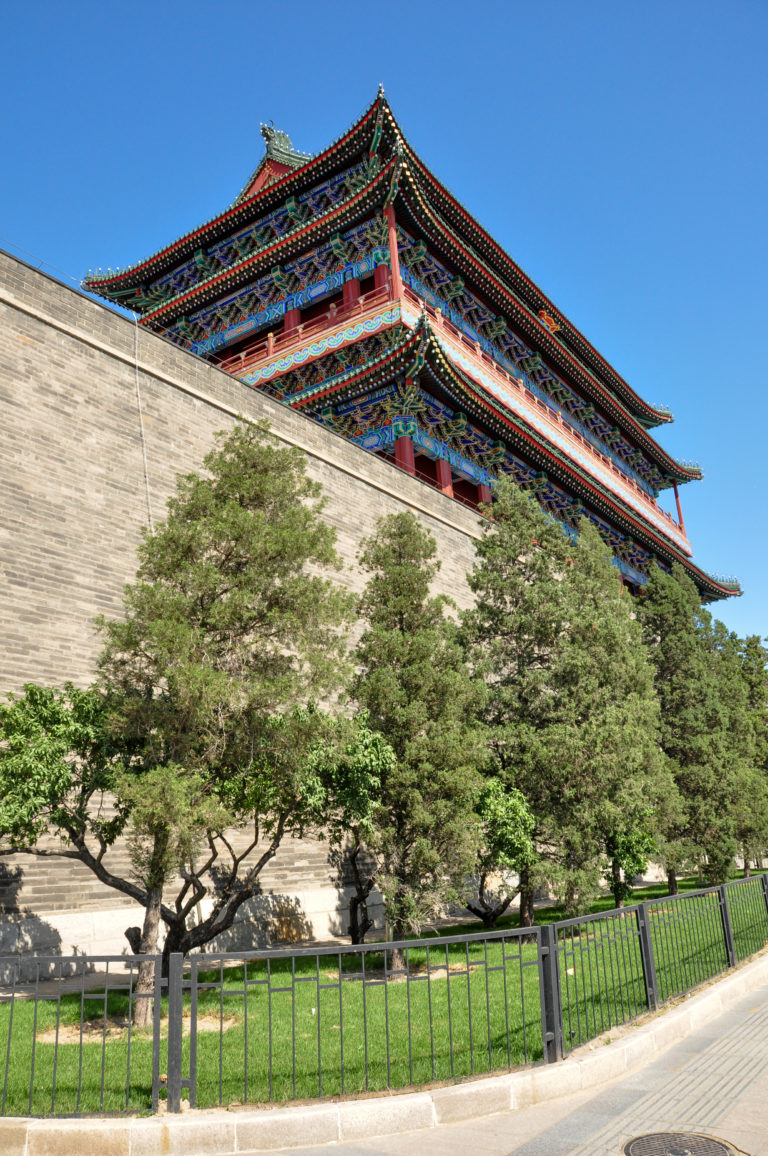 Upper roofline and gray-brick wall of the multi-tiered Zhengyangmen Gate tower seen above trees.