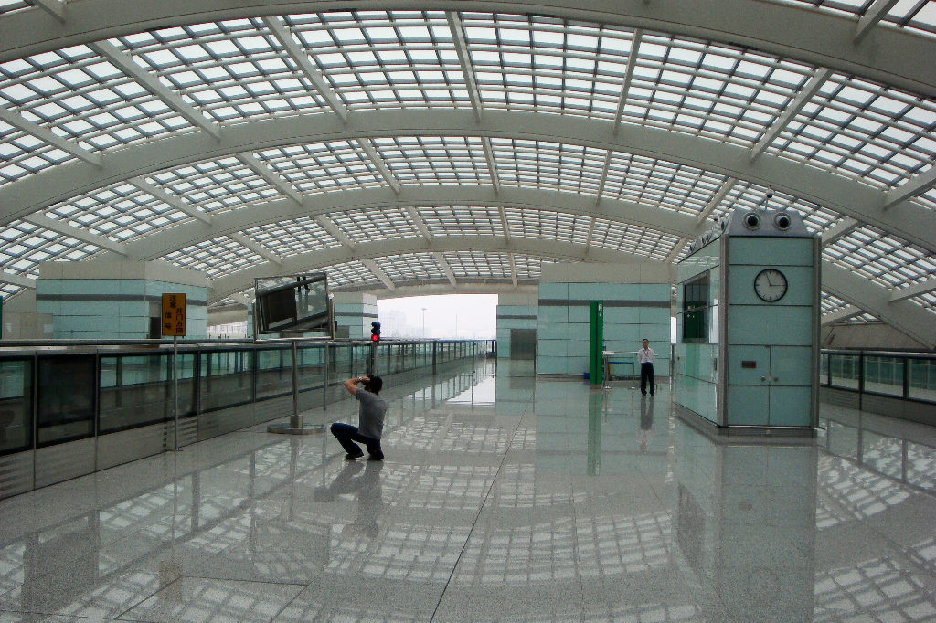 Wide interior view of Beijing Capital International Airport Terminal 3 concourse with moving walkways, a central clock tower, suspended information screen, and a sweeping steel-and-glass roof reflected in the polished floor.