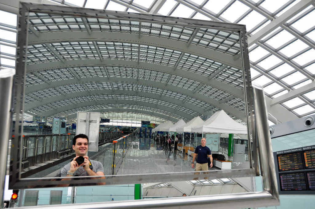 Smiling traveler taking an environmental self-portrait in a large mirror at Beijing Capital International Airport Terminal 3, with the arched roof, concourse, passengers, and flight information displays reflected behind him.
