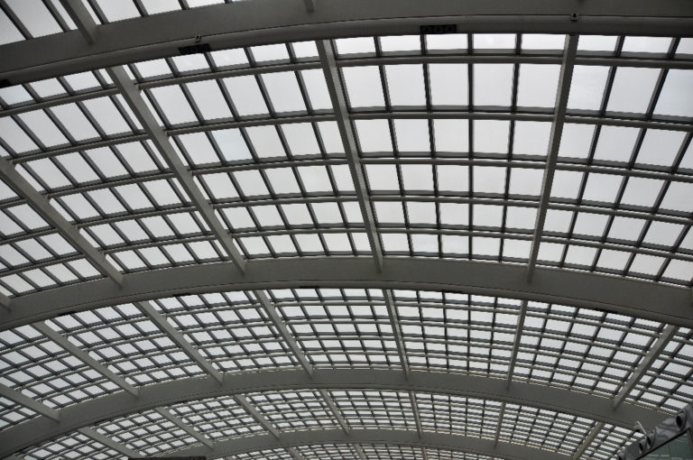 Upward view of the steel-and-glass vaulted roof at Beijing Capital International Airport Terminal 3, showing curved steel ribs and a dense rectangular glazing grid admitting diffuse daylight.