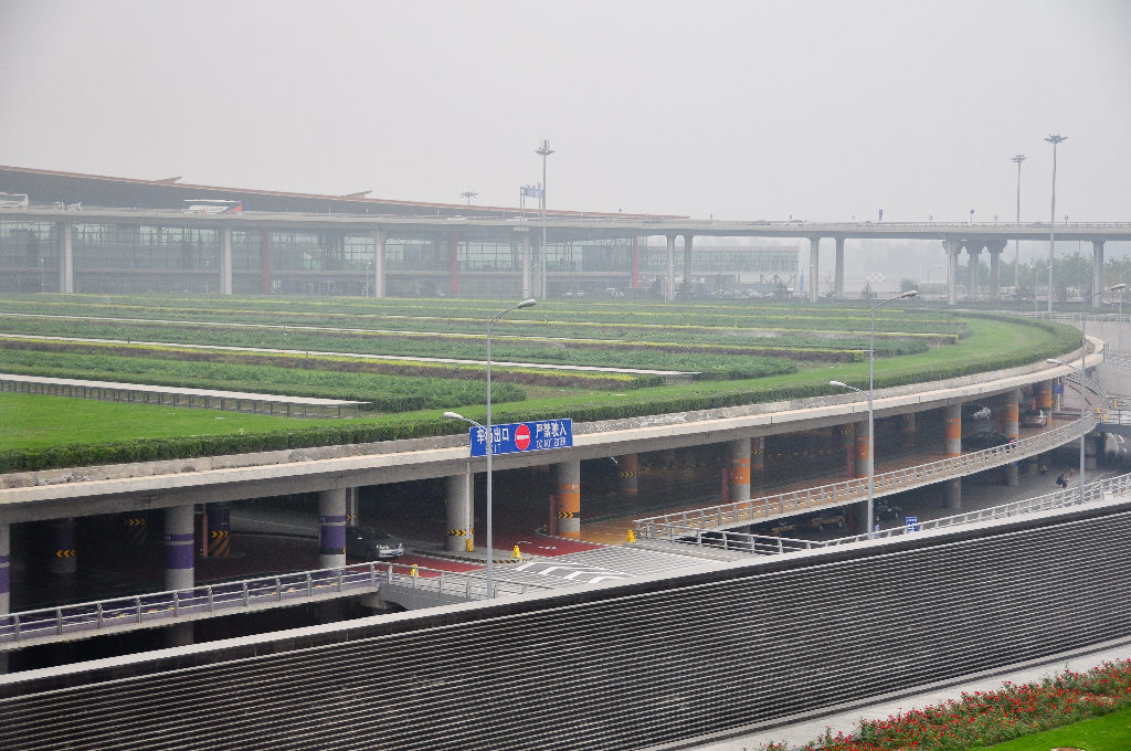 Wide elevated view of curving multi-level access roads and landscaped medians leading to terminal buildings at Beijing Capital International Airport under hazy skies.