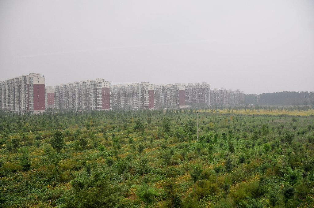 Wide view of a planted greenbelt with young trees near Beijing Capital International Airport, leading toward long rows of multi-story apartment buildings under an overcast sky.