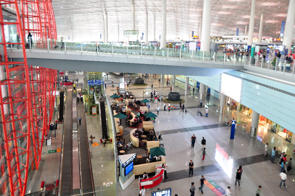 Wide elevated view of the multi-level passenger hall inside Beijing Capital International Airport Terminal 3, showing long escalators, a glass-railed mezzanine, passengers, and airport retail areas on a polished floor.