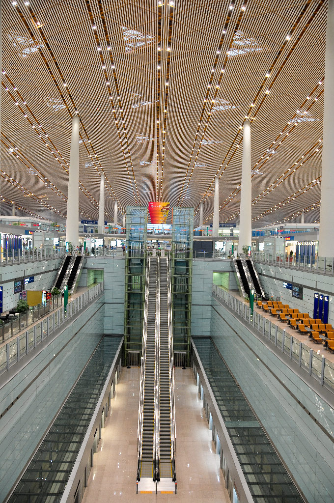 Wide, symmetrical view of the central escalators and glass elevator core in Beijing Capital International Airport Terminal 3, with tall white columns, linear ceiling lights, orange seating on the right, and a red feature in the distance.
