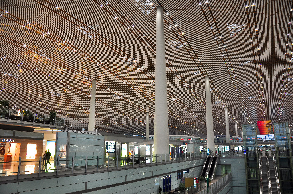 Wide upper-level interior view of Beijing Capital International Airport Terminal 3, showing tall white columns, a dark slatted ceiling with parallel lights, glass balustrades, retail storefronts below, and a red decorative feature in the distance.