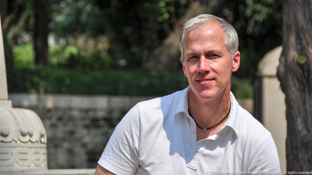 Gray-haired male visitor in a white shirt looking at the camera with stone architecture and greenery of the Giant Wild Goose Pagoda complex in Xi'an behind him.