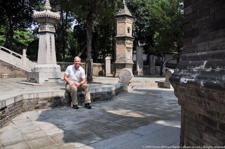 Male visitor seated on a low stone ledge beside carved stone ornaments and upright markers in a paved courtyard at Da Ci'en Temple in Xi'an.