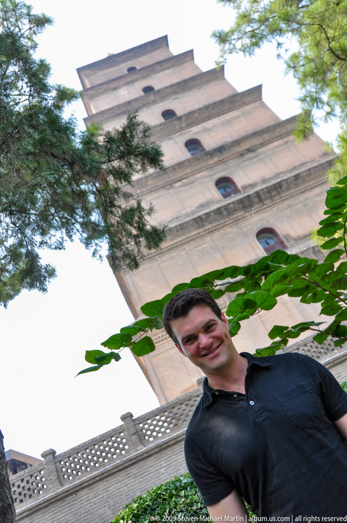 Smiling male visitor in the foreground with the Giant Wild Goose Pagoda rising behind him, framed by green leaves, seen from a low angle.