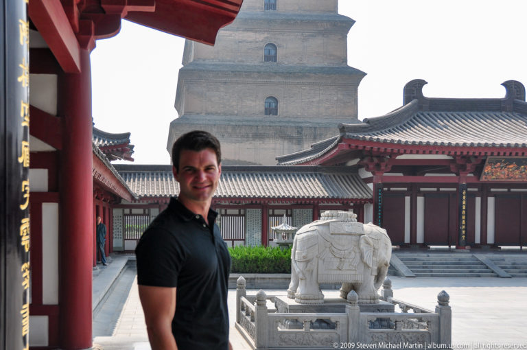 Male visitor standing beside a white stone elephant sculpture in a red-columned courtyard with the Giant Wild Goose Pagoda in the background.
