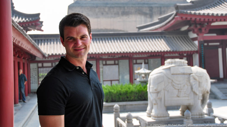 Male visitor standing beside a white stone elephant sculpture in a red-columned courtyard with the Giant Wild Goose Pagoda in the background.