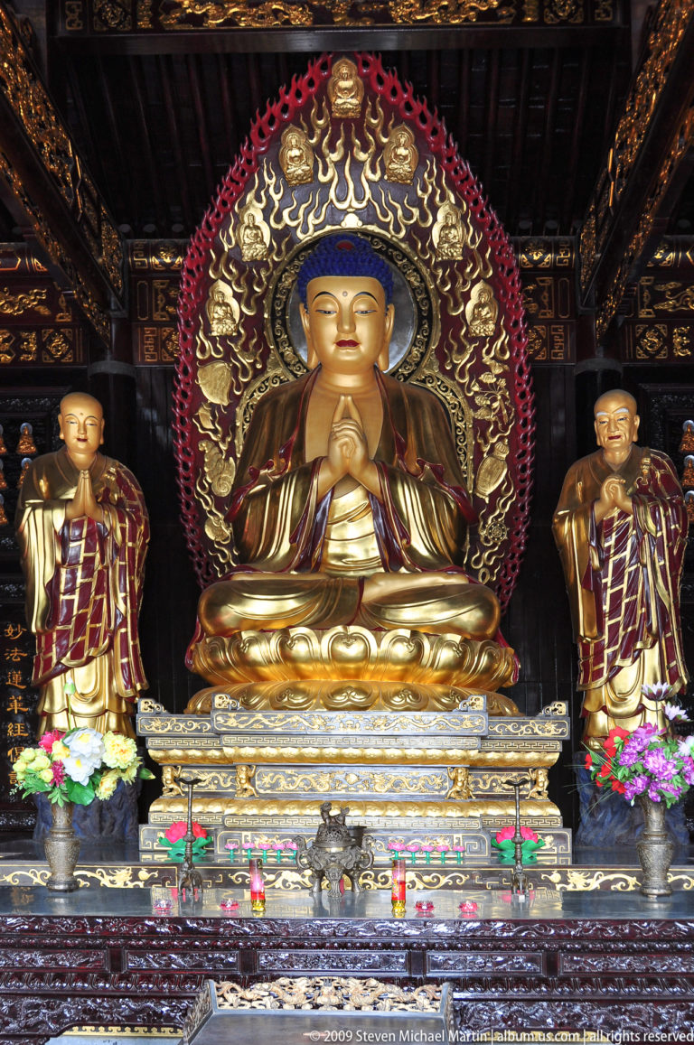 Gilded seated Buddha on a lotus pedestal with ornate halo, flanked by two standing attendant figures in a temple setting.