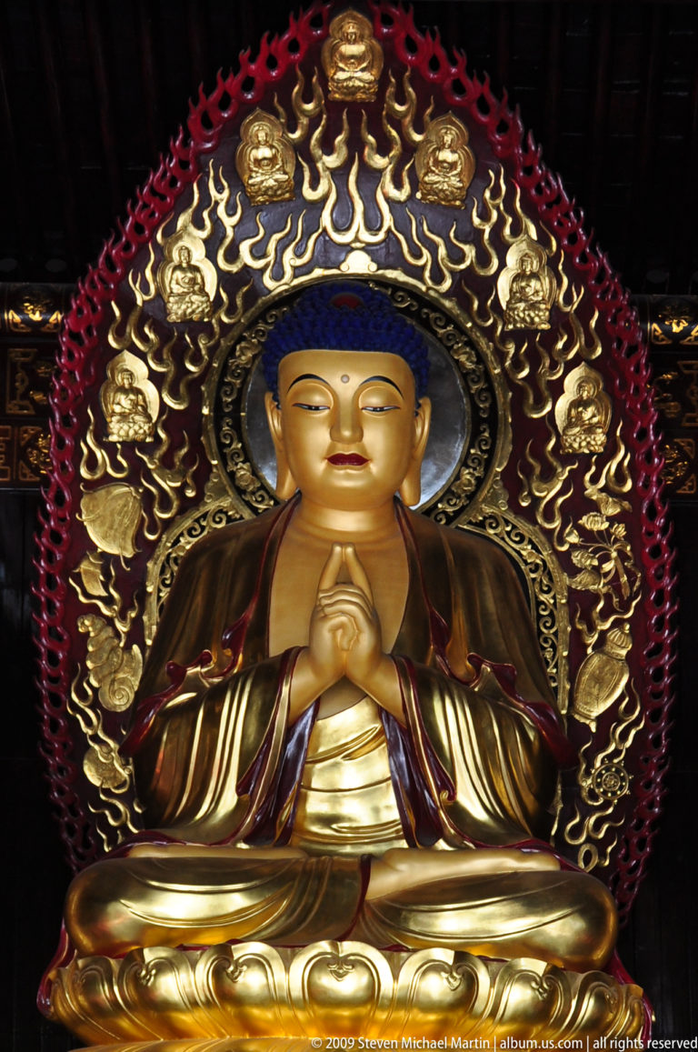 Close frontal view of a gilded Buddha statue with blue hair curls, elongated earlobes, and a precise teaching mudra, framed by an ornate circular halo.