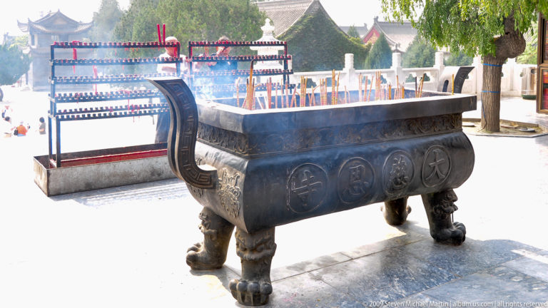 Large bronze incense burner on animal-form legs with many upright burning incense sticks emitting smoke.