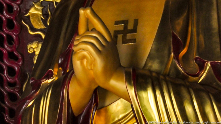 Gilded Buddha hands in a ritual mudra beneath a Buddhist swastika symbol, framed by dark and gold robe folds.