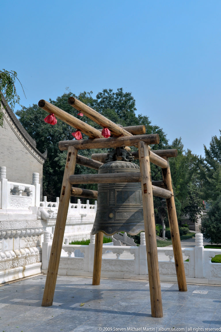 Large bronze temple bell with decorative panels hanging from a timber frame in front of white stone balustrades.