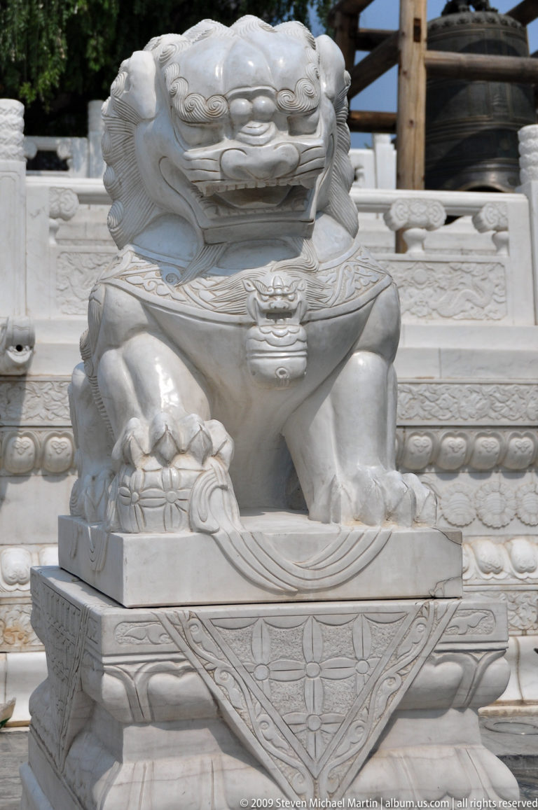 Close view of a white stone guardian lion’s paw resting on a rounded decorative element, with a small lion-head pendant on its chest and patterned stonework behind.