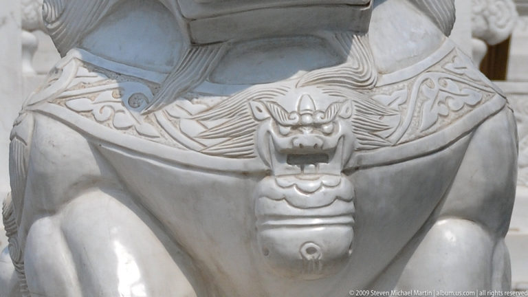 Close-up of a small lion-head pendant hanging from the collar on a stone guardian lion’s chest.