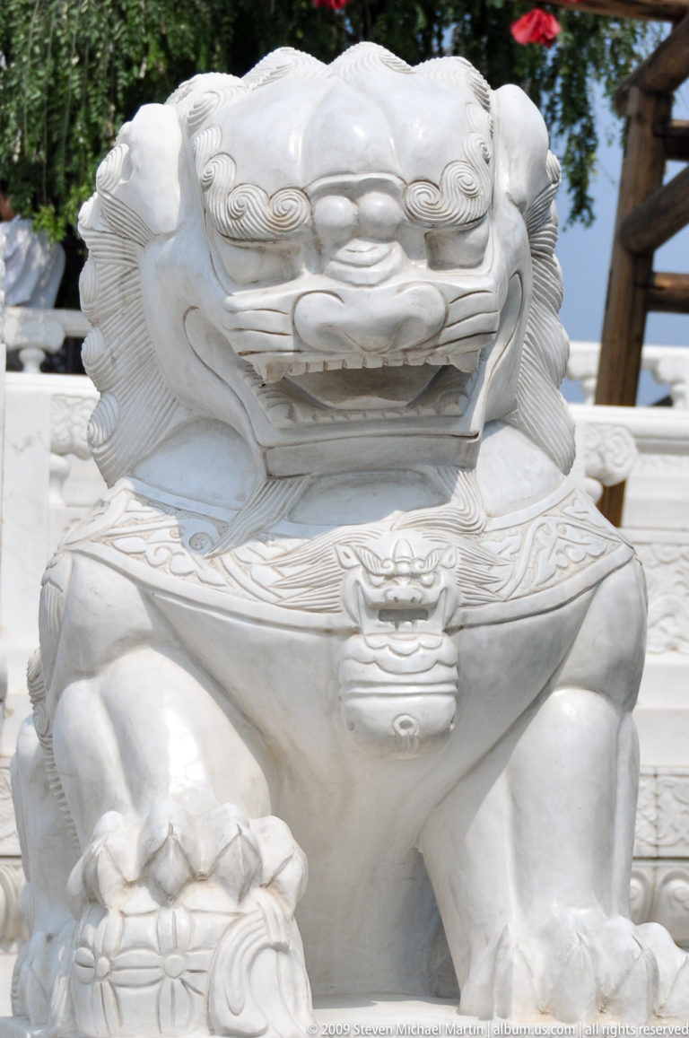 Close-up of the upper torso of a white stone guardian lion showing its open mouth, carved mane, and pendant-like lion-head chest ornament.