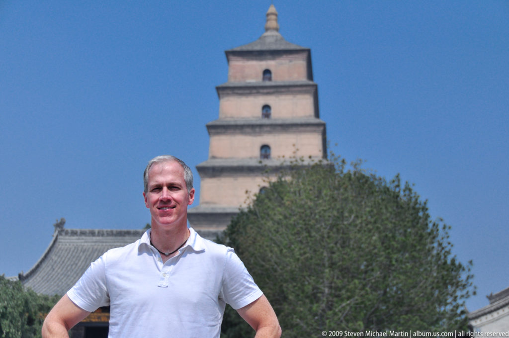 Smiling male visitor standing in front of the seven-tiered Giant Wild Goose Pagoda rising above trees and traditional tiled roofs in Xi'an.