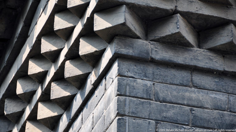 Geometric projecting brick pattern in gray masonry on a Da Ci'en Temple wall, forming a textured band below the roof.