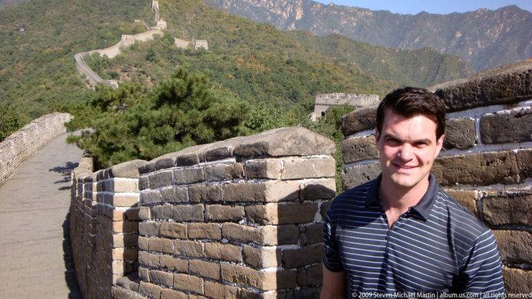 Smiling male visitor standing beside a brick parapet on the Great Wall of China, with watchtowers and wooded mountains in the background.