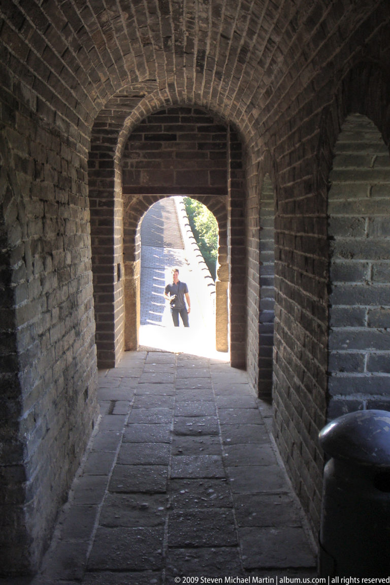 View from inside a dark Great Wall watchtower, looking through a pointed brick archway to a sunlit rampart where a visitor stands in silhouette.