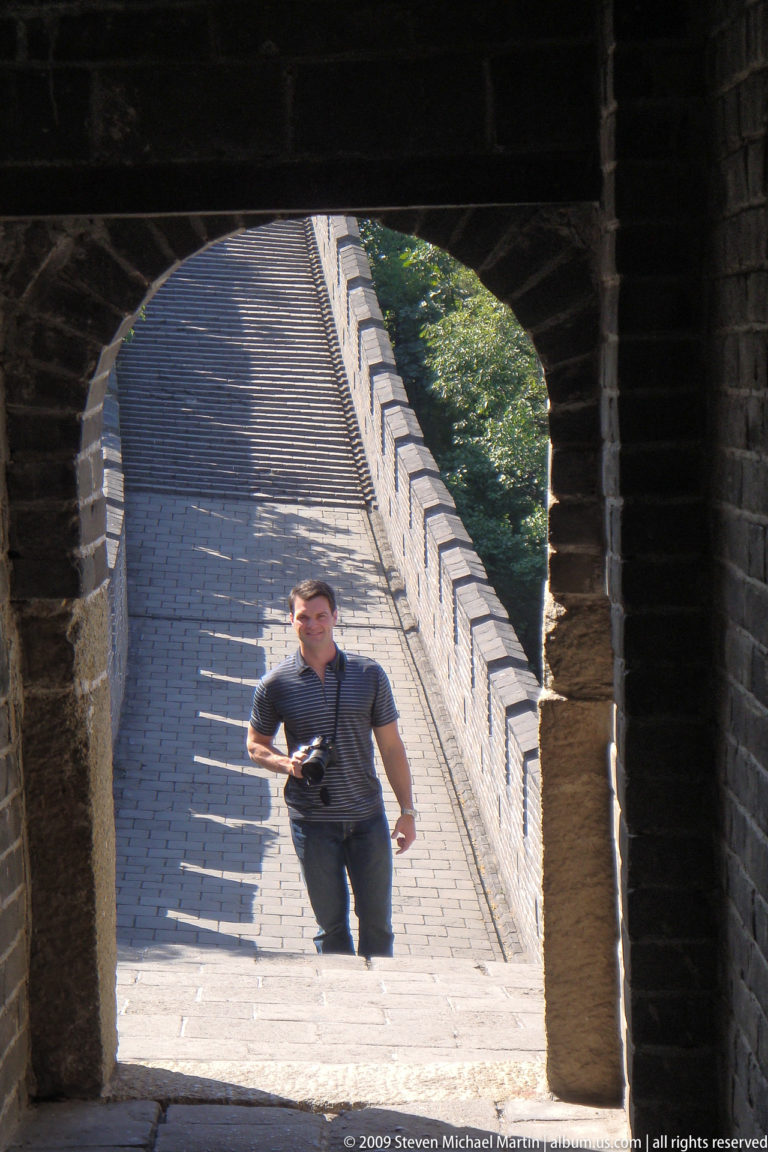 Male visitor holding a camera in the arched doorway of a Great Wall watchtower with steep stairs rising behind him.