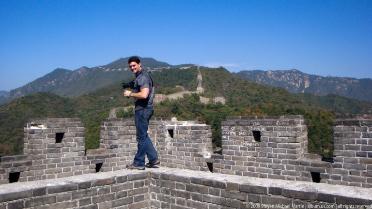 Male visitor standing on a raised brick battlement of the Great Wall with crenellations and mountains in the background.