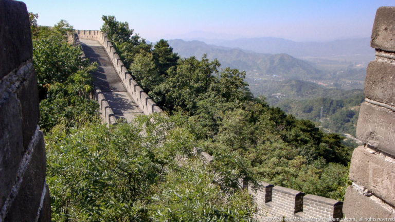 Steep brick-paved Great Wall rampart with crenellated parapets climbing a wooded ridge at Mutianyu, with hazy mountains in the distance.
