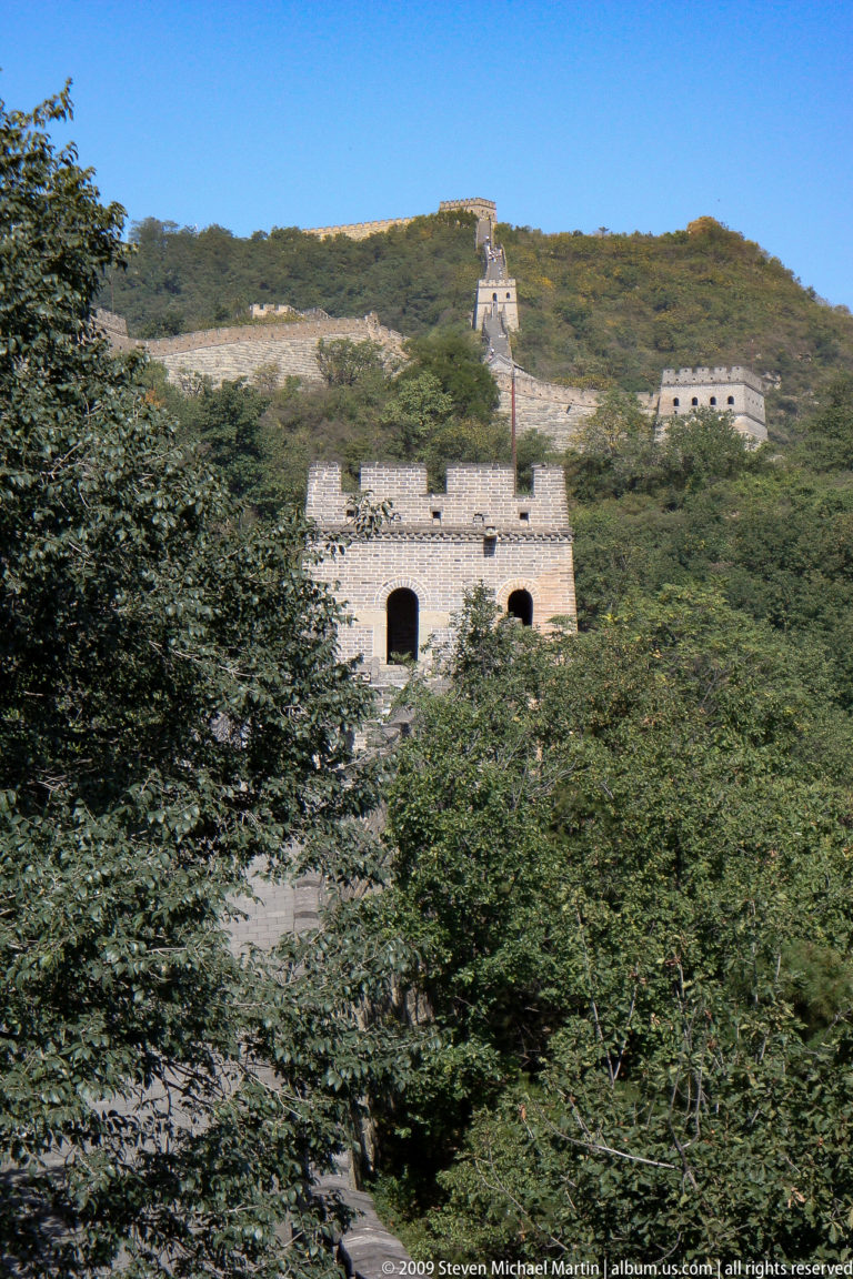 Telephoto view of a square Great Wall watchtower with two tall arched openings and a crenellated roofline, partly obscured by surrounding trees.
