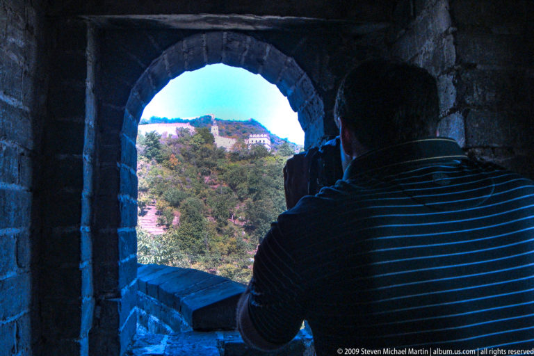 Silhouetted visitor inside a dark Great Wall watchtower looking through an arched opening toward a distant sunlit tower and wall on a ridge.