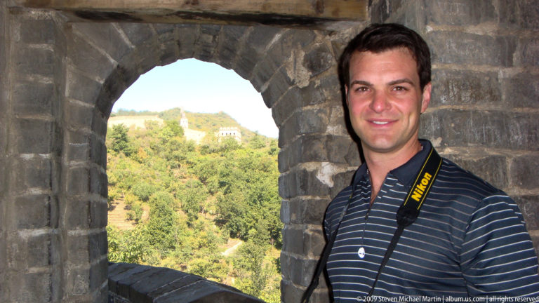 Male visitor with a camera strap standing in the arched opening of a Great Wall watchtower, with distant wall towers visible through the opening.