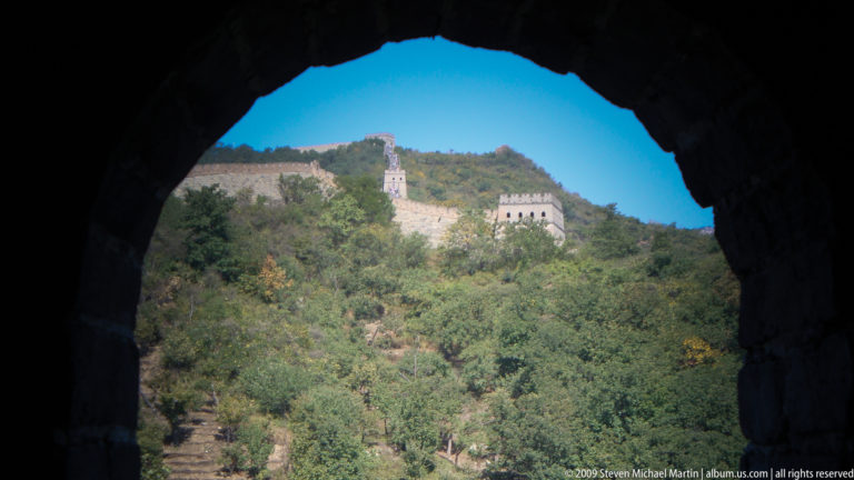 View through a dark watchtower arch at Mutianyu toward sunlit Great Wall towers and crenellated walls running along the opposite wooded ridge.