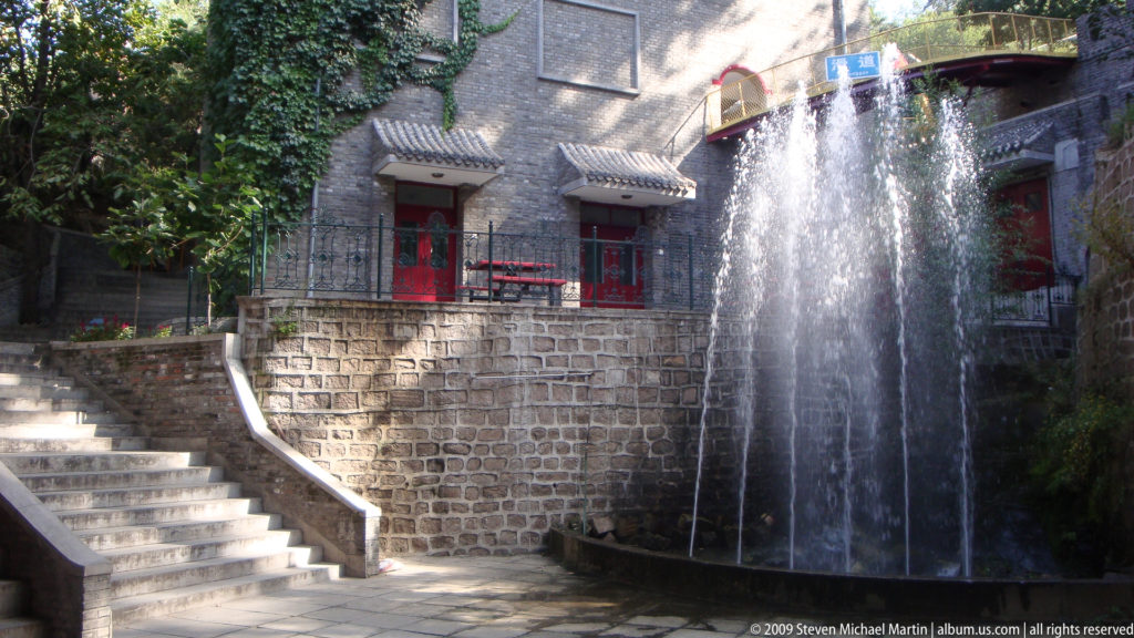 Circular waterfall fountain in front of a gray-brick visitor building with red doors at the Mutianyu Great Wall tourist complex.