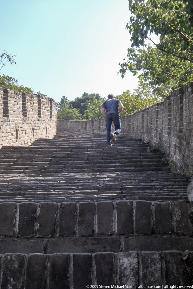 Male visitor climbing a steep, irregular stone stairway between brick parapets on the Great Wall at Mutianyu.