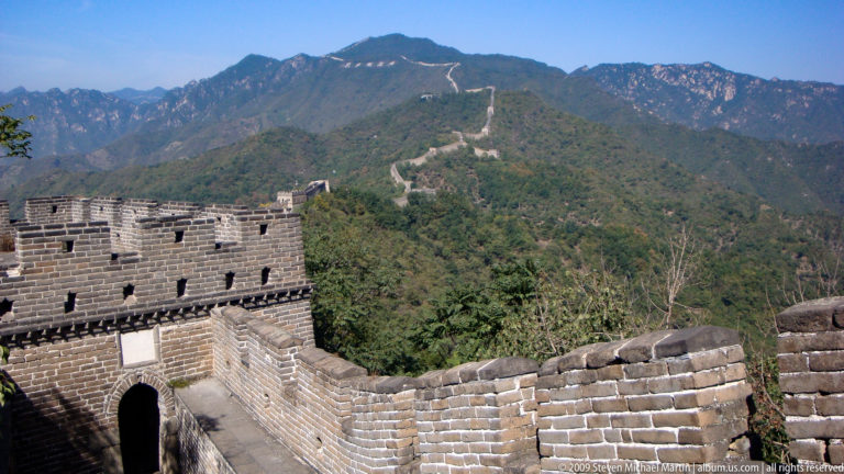 Ming-era Great Wall watchtower and rampart at Mutianyu extending along a wooded mountain ridge with multiple distant towers visible.